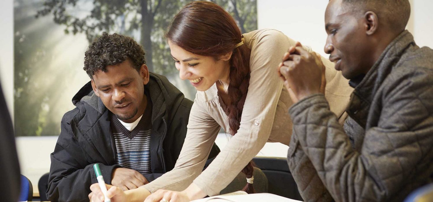 A photograph showing a woman helping two men with English language lessons.